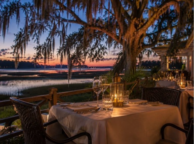 Romantic waterfront restaurant patio on Hilton Head Island at sunset, featuring candlelit tables, Spanish moss-draped oak trees, and scenic Lowcountry marsh views.