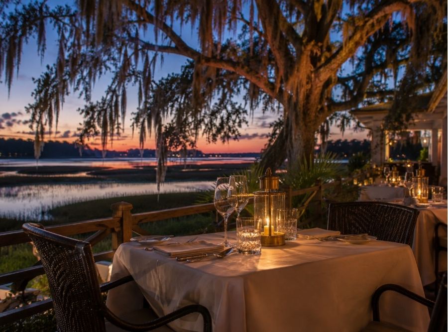 Romantic waterfront restaurant patio on Hilton Head Island at sunset, featuring candlelit tables, Spanish moss-draped oak trees, and scenic Lowcountry marsh views.